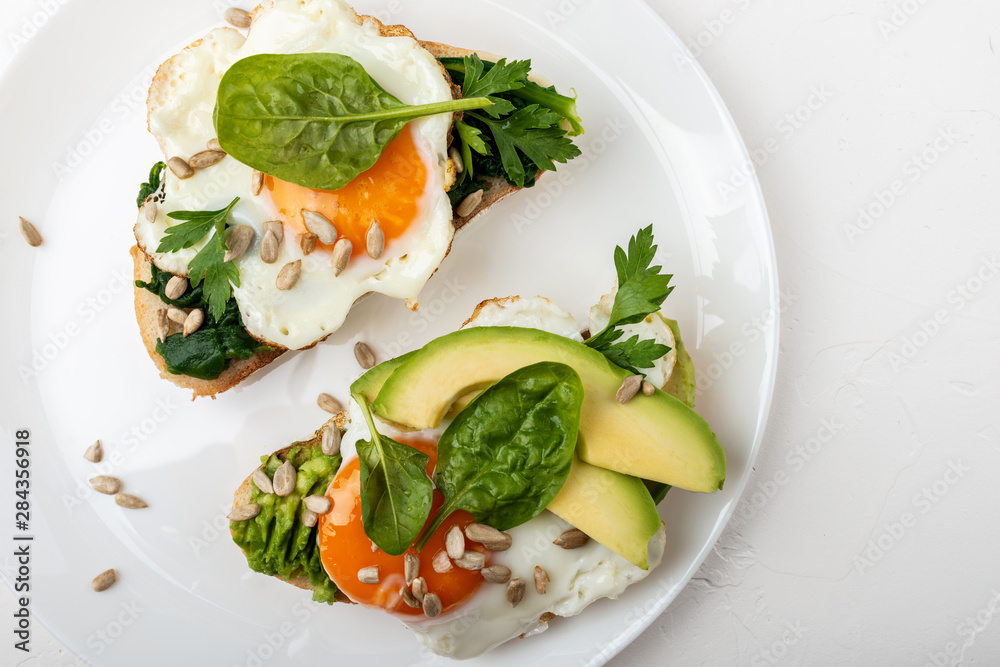 Fried eggs on a toasts bread with avocado, spinach and seeds on a white plate on the white background.