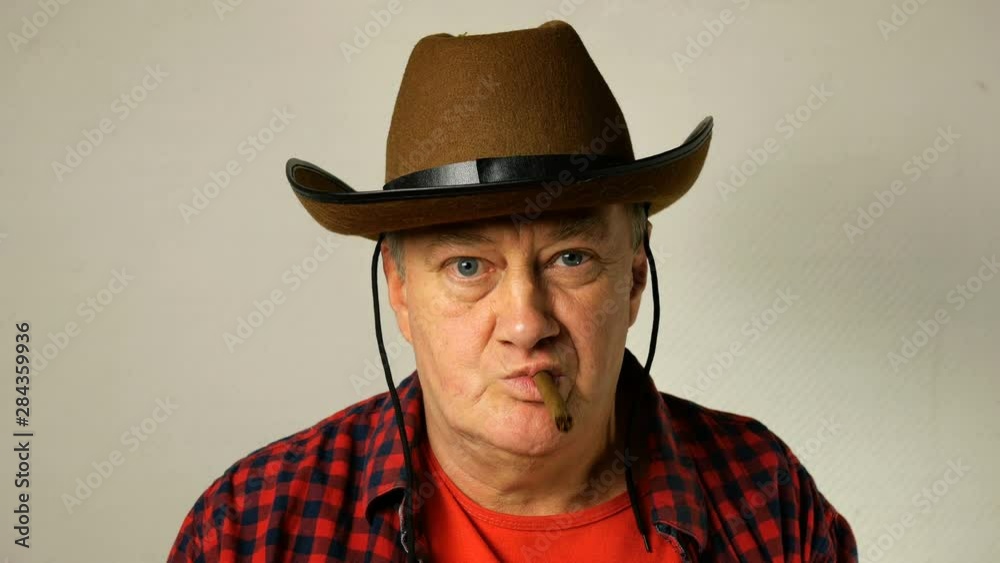 Senior man of caucasian ethnicity in a cowboy hat with a cheeky look, smokes a cigar and blows smoke towards the camera. Close-up. Senior man of caucasian ethnicity in a cowboy hat with a cheeky look, smokes a cigar and blows smoke towards the camera. Close-up.