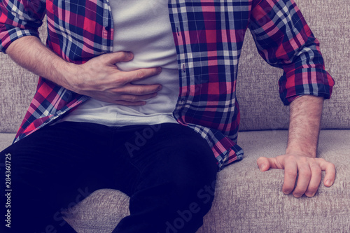 Man suffering stomach ache, clutching his stomach from acute pain, sitting on a couch in home, man's hands, close up