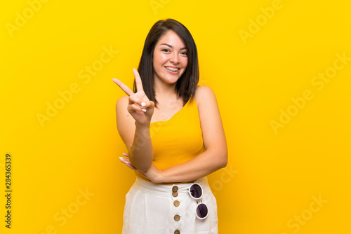 Young Mexican woman over isolated yellow background smiling and showing victory sign