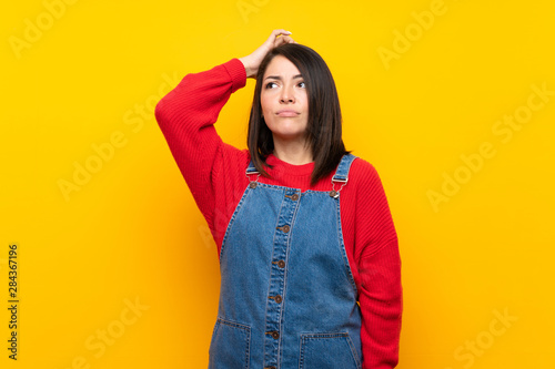 Young Mexican woman with overalls over yellow wall having doubts while scratching head
