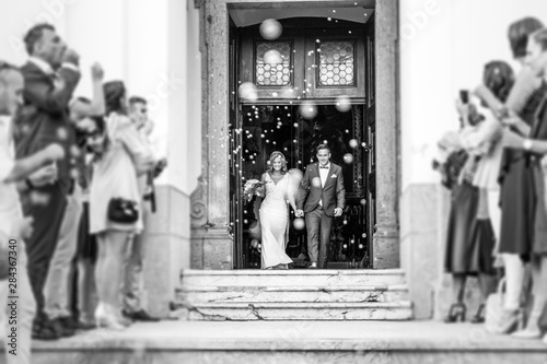 Newlyweds exiting the church after the wedding ceremony, family and friends celebrating their love with the shower of soap bubbles, custom undermining traditional rice bath. Black and white.
