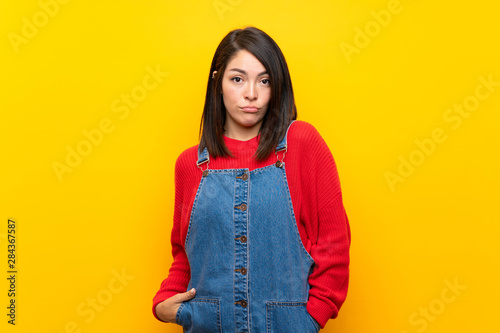Young Mexican woman with overalls over yellow wall with sad and depressed expression © luismolinero