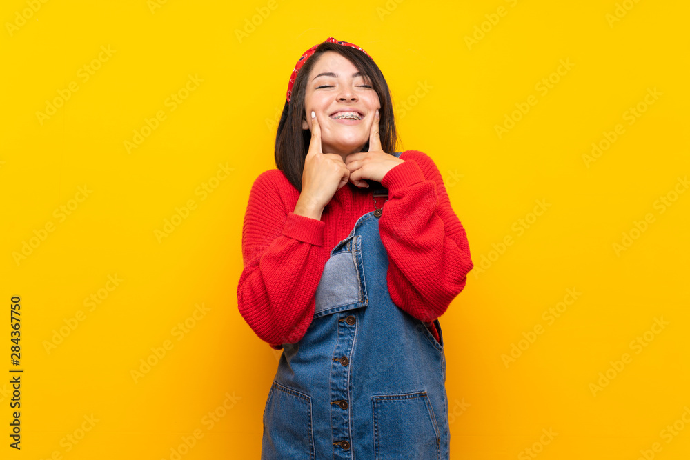 Young Mexican woman with overalls over yellow wall smiling with a happy and pleasant expression Young Mexican woman with overalls over yellow wall smiling with a happy and pleasant expression