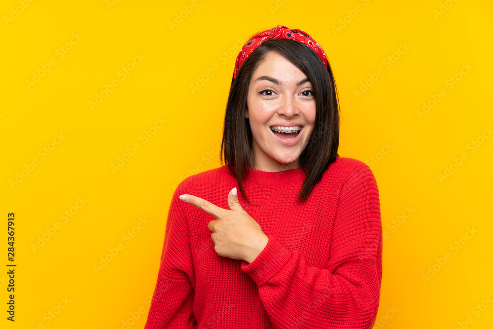 Young Mexican woman with red sweater over yellow wall pointing finger to the side Young Mexican woman with red sweater over yellow wall pointing finger to the side