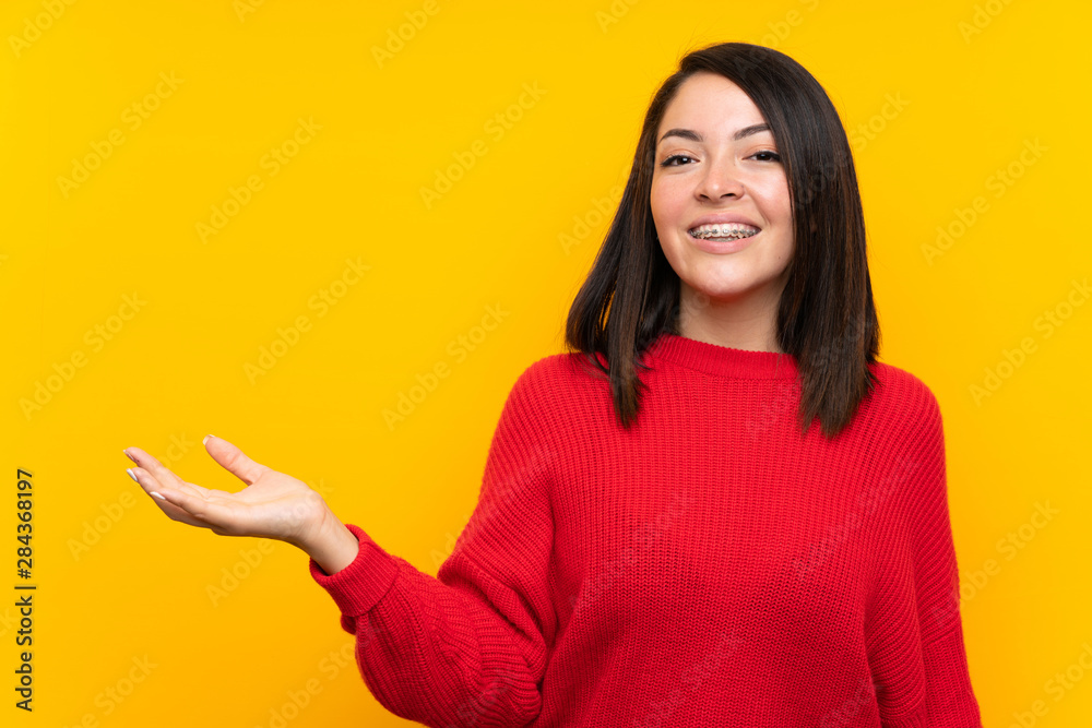 Young Mexican woman with red sweater over yellow wall holding copyspace imaginary on the palm Young Mexican woman with red sweater over yellow wall holding copyspace imaginary on the palm