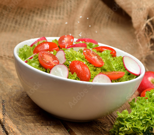 Close-up of fresh green salad Lollo Biondo with wet tomatoes and radishes with drops in a white bowl on wooden table
