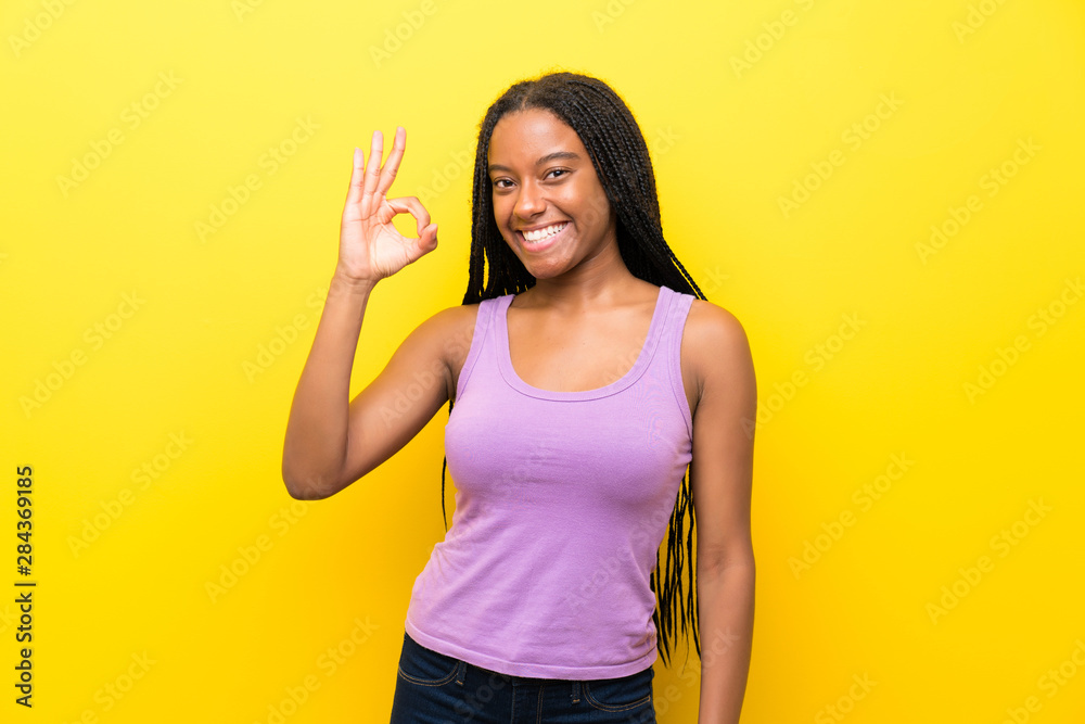 African American teenager girl with long braided hair over isolated yellow wall showing ok sign with fingers African American teenager girl with long braided hair over isolated yellow wall showing ok sign with fingers