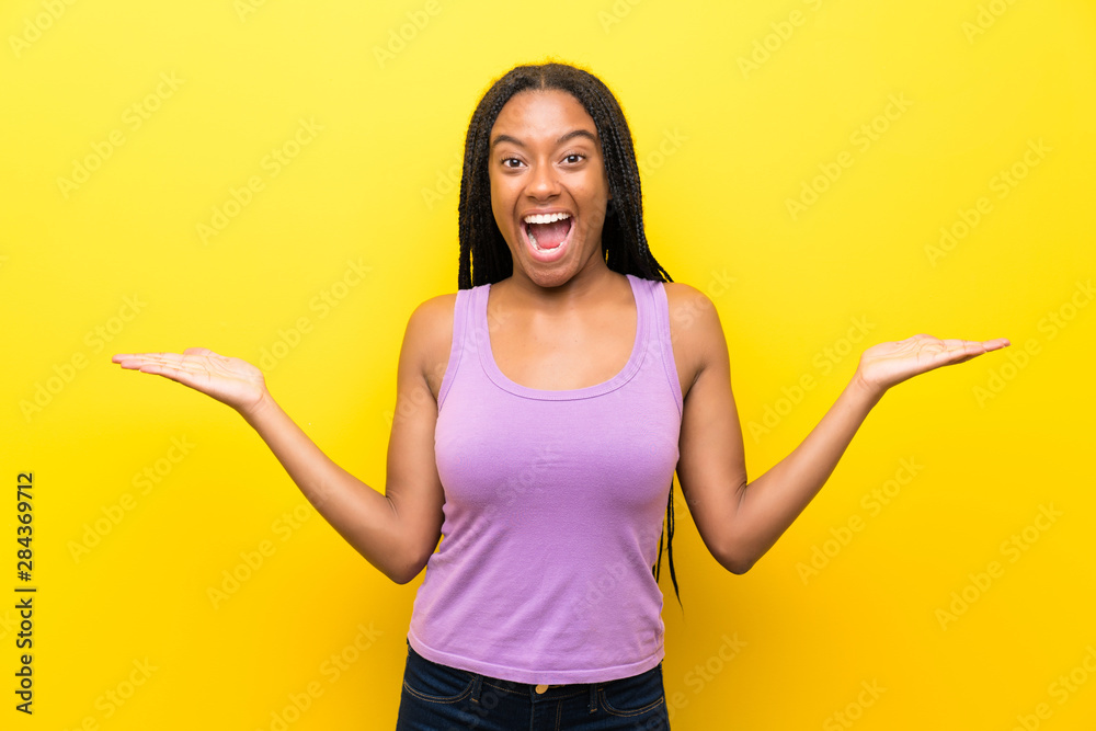 African American teenager girl with long braided hair over isolated yellow wall with shocked facial expression African American teenager girl with long braided hair over isolated yellow wall with shocked facial expression