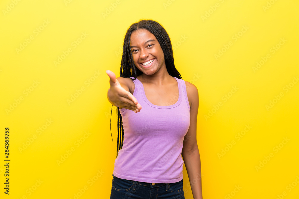 African American teenager girl with long braided hair over isolated yellow wall shaking hands for closing a good deal African American teenager girl with long braided hair over isolated yellow wall shaking hands for closing a good deal