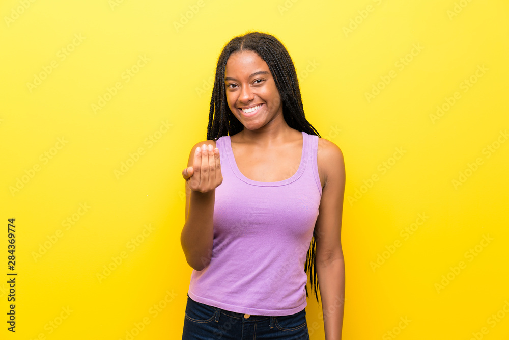 African American teenager girl with long braided hair over isolated yellow wall inviting to come with hand. Happy that you came African American teenager girl with long braided hair over isolated yellow wall inviting to come with hand. Happy that you came
