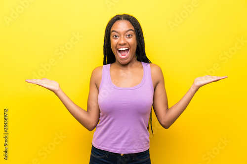 African American teenager girl with long braided hair over isolated yellow wall with shocked facial expression
