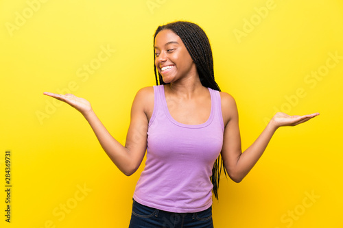 African American teenager girl with long braided hair over isolated yellow wall holding copyspace with two hands