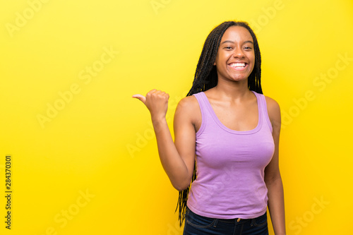 African American teenager girl with long braided hair over isolated yellow wall pointing to the side to present a product