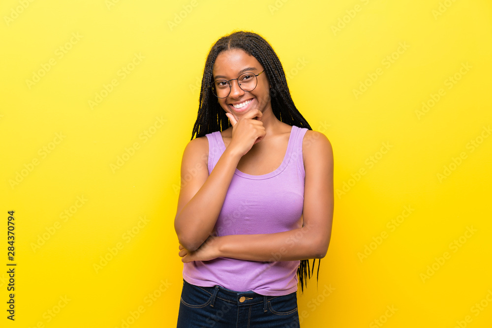 African American teenager girl with long braided hair over isolated yellow wall with glasses and smiling African American teenager girl with long braided hair over isolated yellow wall with glasses and smiling
