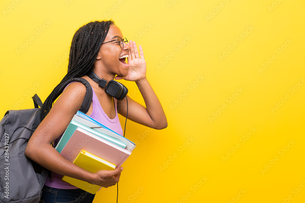 African American teenager student girl with long braided hair over isolated yellow wall shouting with mouth wide open - obrazy, fototapety, plakaty 