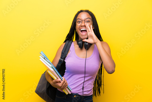 African American teenager student girl with long braided hair over isolated yellow wall shouting with mouth wide open