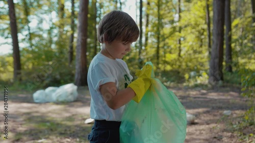 Face boy voluteers activists child in gloves tidying up rubbish in park or forest look at camera smile save environmentplastic pollution bag bottle recycle ecology garbage nature care slow motion photo