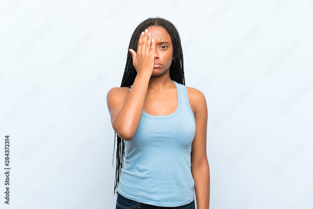 African American teenager girl with long braided hair over isolated blue background covering a eye by hand