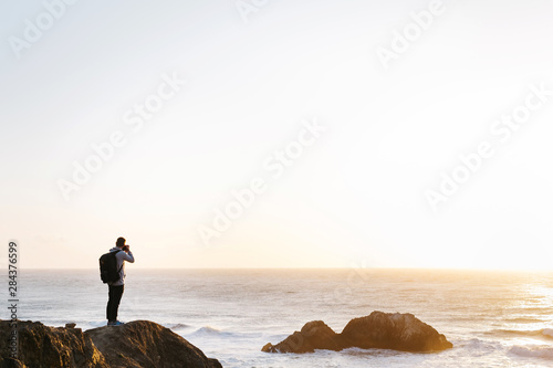 Hipster young man hiker standing on cliffs photographing sunset over ocean