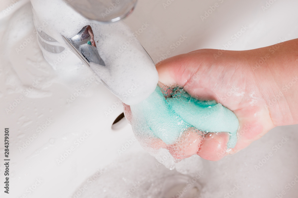 Fototapeta premium Maid holding sponge under running water. Bubbles forming in sink. Shiny and clean bathroom.