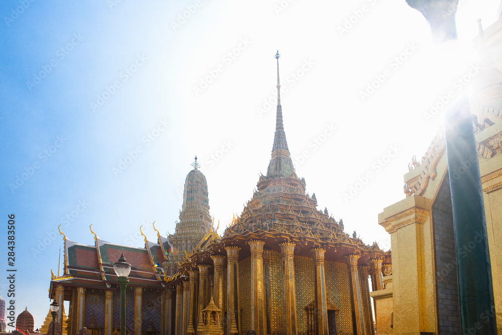 Fototapeta premium Bangkok, Thailand - A golden Buddhist temple is surrounded by columns supporting a decorative roof. A sunburst can be seen in the upper right of the shot.