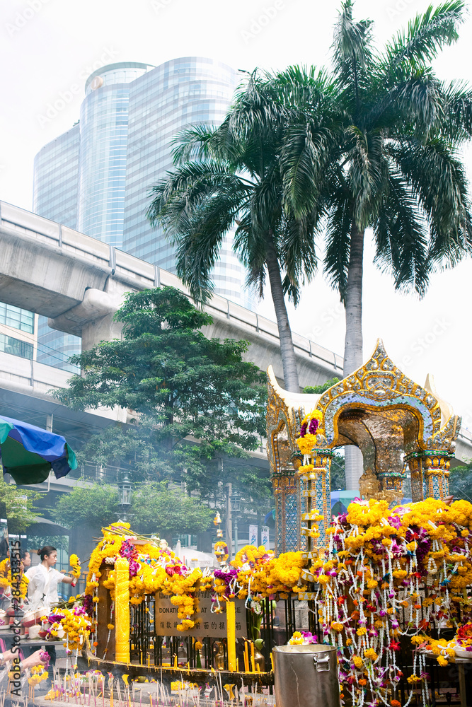Bangkok, Thailand - A shrine with incense, flowers and offerings is set outside in a city setting. A skyscraper can be seen in the background.