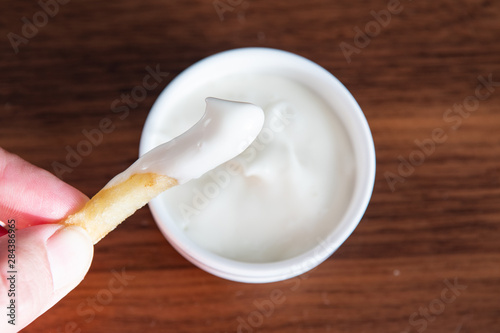 Hand holding one piece of french fries with in mayonnaise and sauce in the background. Mayonnaise in small round white ceramic dish on wooden table.