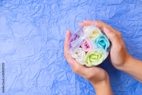 Rose flowers shaped scented soap petals in transperent round box on young girl's hands on blue crimpled background. Handcrafted soap petals. photo