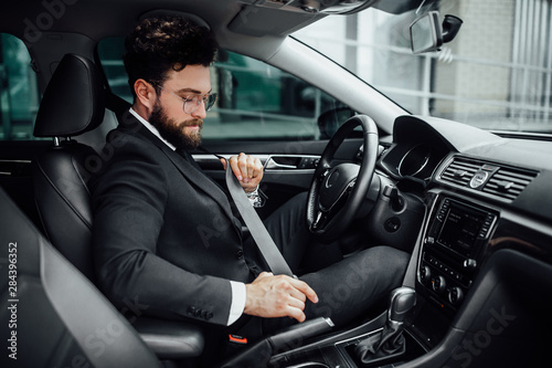 The young handsome bearded man fastening seat belt in a black business suit. A driver in a new car. © Тарас Нагирняк