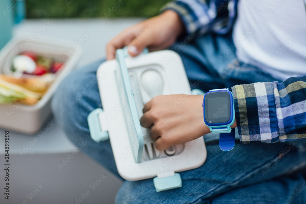 Close up photo, lunch boxes with healthy food and child boy with smartwatches on hands.