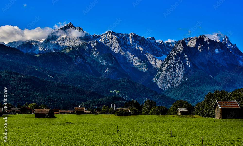 Garmisch Partenkirchen, Germany. View of the Alps with snow, blue sky, and clouds, with a green pasture and brown chalet-style buildings