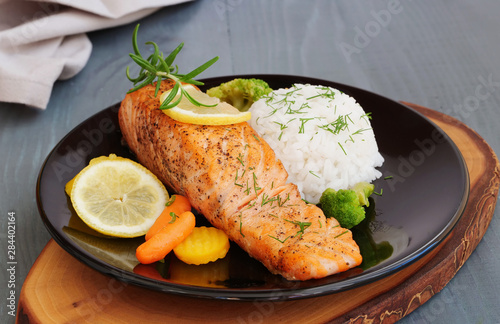 Grilled salmon fillet with rice and vegetables on black plate over wooden table