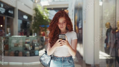 Extremely beautiful cute young redheaded student girl walking down the shopping center while actively texting via her smartphone and smiling. Modern communication, having fun, shopping time. Female