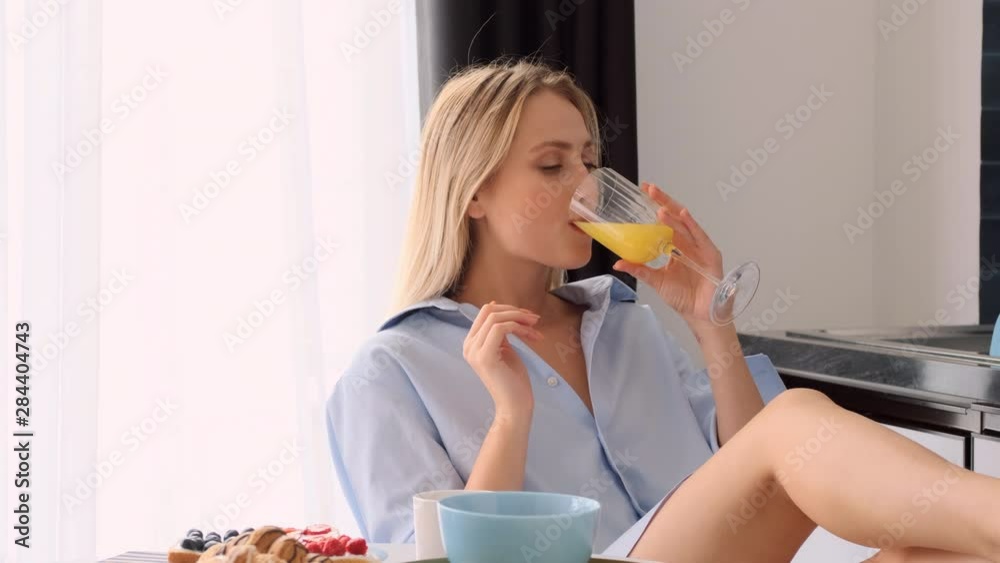 Tracking out shot of beautiful smiling blond woman in male shirt eating croissant enjoying the taste and drinking orange juice on modern kitchen Tracking out shot of beautiful smiling blond woman in male shirt eating croissant enjoying the taste and drinking orange juice on modern kitchen