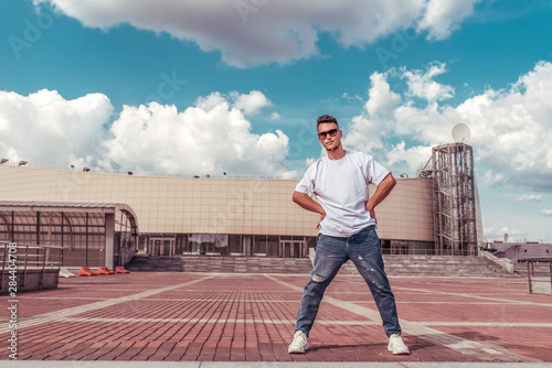 young guy a dancer in a white T-shirt, jeans, posing for a leader, in summer in city, in sunglasses, background building clouds, active hip hop, youth lifestyle, free space for text © byswat