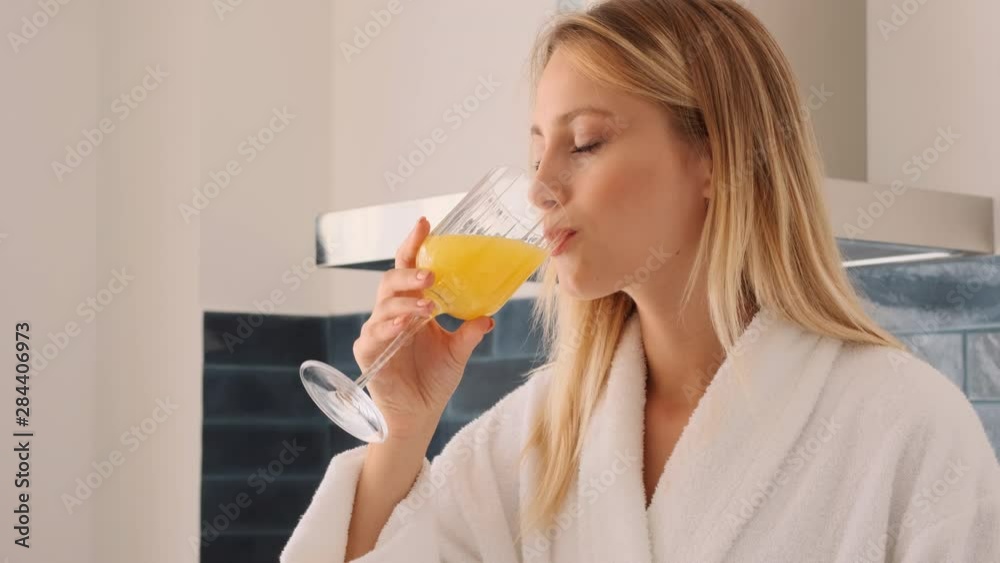 Tracking out shot of beautiful blond woman in bathrobe drinking orange juice and happily using cellphone on kitchen at home Tracking out shot of beautiful blond woman in bathrobe drinking orange juice and happily using cellphone on kitchen at home