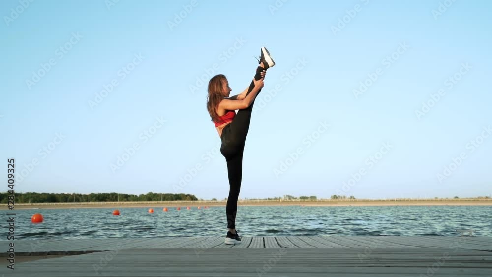 A young athletic Caucasian girl in a red tank top and leggings shows her stretch and flexibility against the backdrop of a lake. Beautiful dance. Stretching, split.