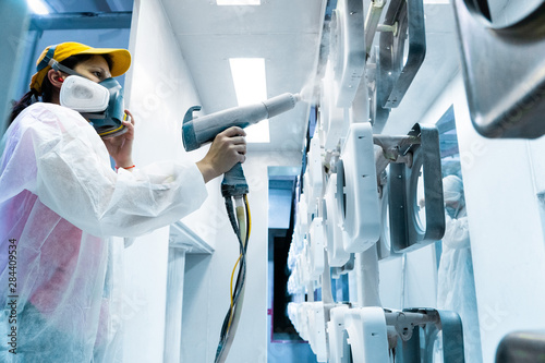 Powder coating of metal parts. A woman in a protective suit sprays white powder paint from a gun on metal products