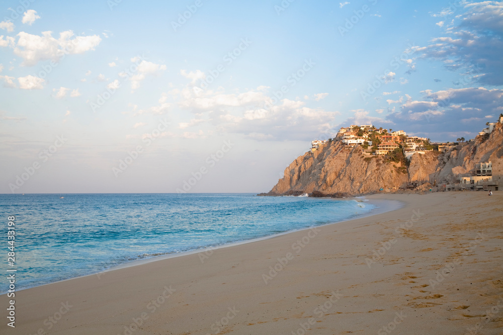 Cabo San Lucas, Baja California Sur, Mexico - A beach with natural rock formation in the distance. Resorts or homes can be seen on the rocks.