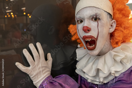The guy in the IT suit opened his mouth. The face is a very scary clown with red hair and sharp teeth close-up. Portrait of a terrible hero with reflection on a dark wall. Cosplay for halloween.
