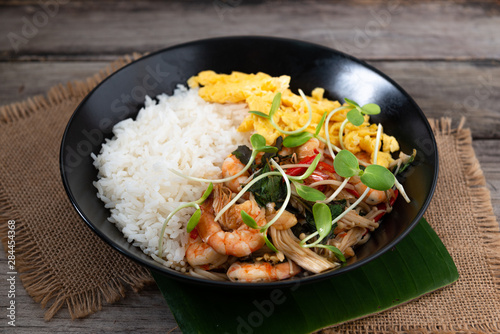 Rice topped with stir-fried shrimp and basil, and fried eggs in white dish.