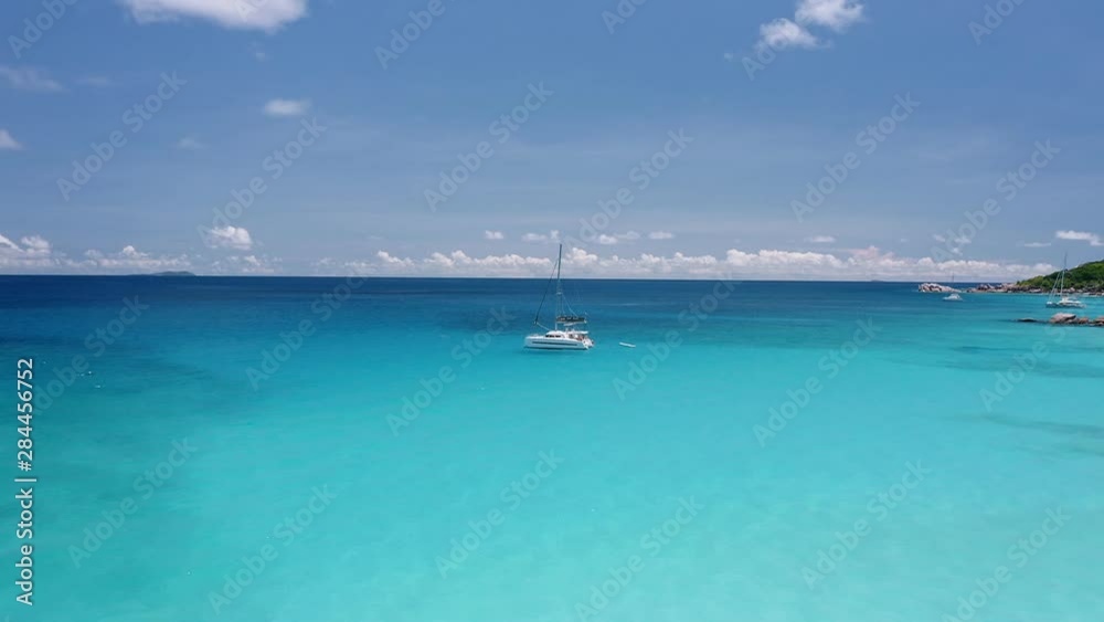 Aerial circle footage of luxury catamaran yacht boat moored in transparent turquoise blue lagoon with white clouds on horizon. La Digue island, Seychelles. Travel concept