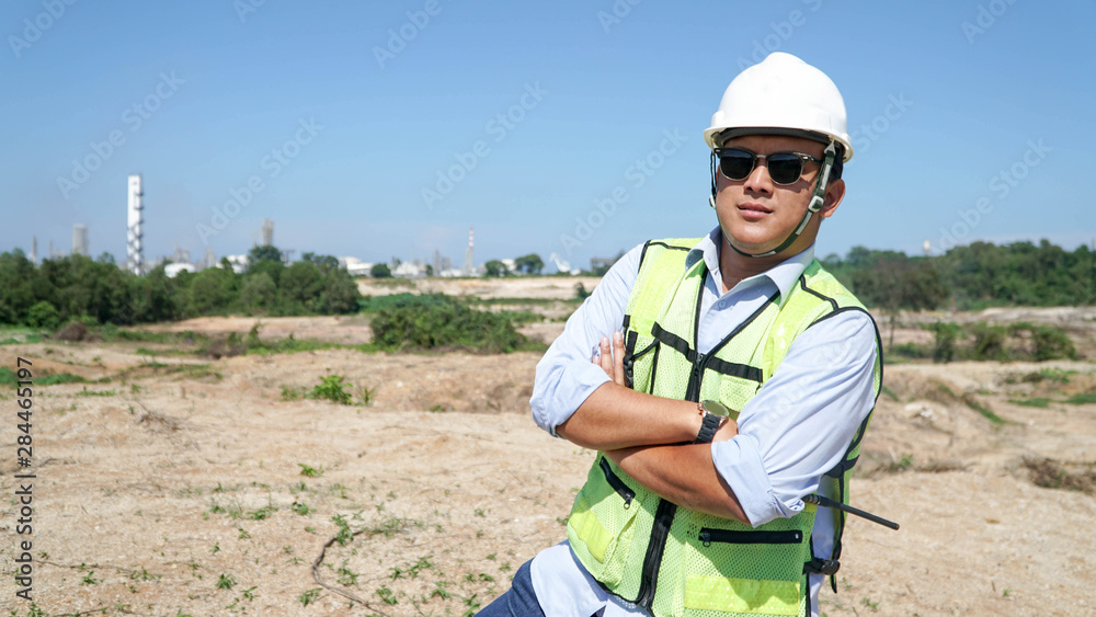 close up front view portrait of male engineer smile at camera while cross his arm on outdoor bright blue sky dress with protective vest, hard helmet and black eye glasses close up front view portrait of male engineer smile at camera while cross his arm on outdoor bright blue sky dress with protective vest, hard helmet and black eye glasses