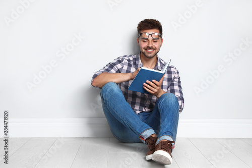 Handsome young man reading book on wooden floor near light wall