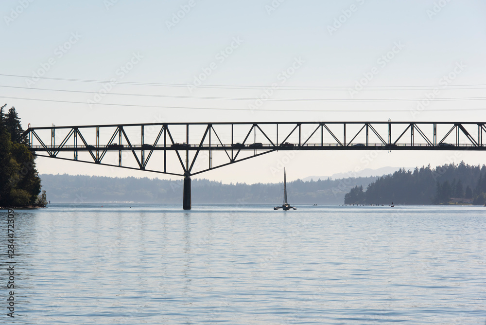 USA, Washington State. Puget Sound. Trimaran passes under Agate Pass Bridge between Kitsap Peninsula and Bainbridge Island. Afternoon traffic silhouetted