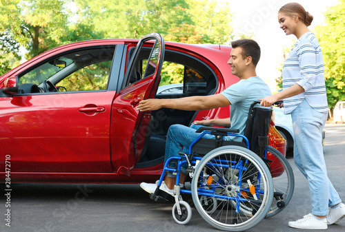 Young woman helping disabled man in wheelchair to get into car outdoors