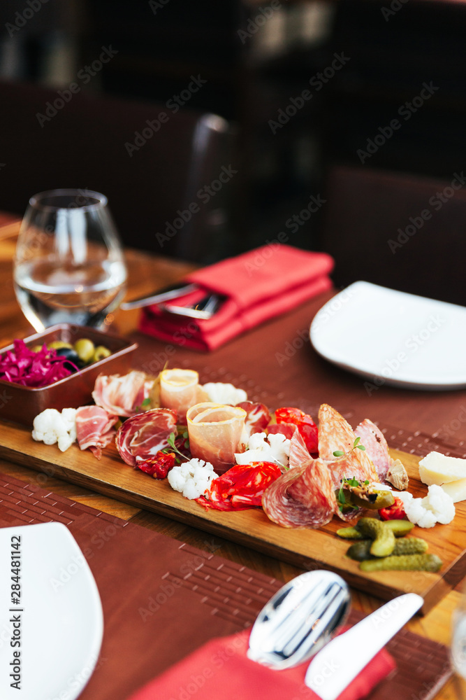 Cold cuts on wooden board with prosciutto, bacon, salami and sausages. Meat platter appetizers served with pickle and olives on dining table with cutlery.