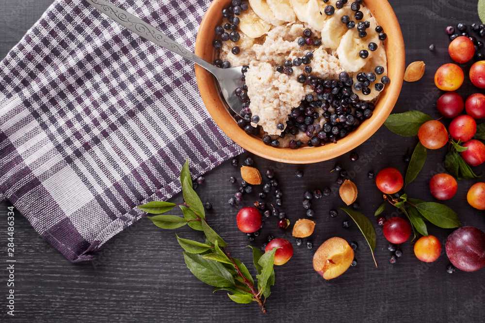 Image of orange plate of oatmeal with blueberries and bananas stands on delicate black wooden background on which lies checkered cotton towel. Healthy eating, breakfast, healthy food and diet concept.
