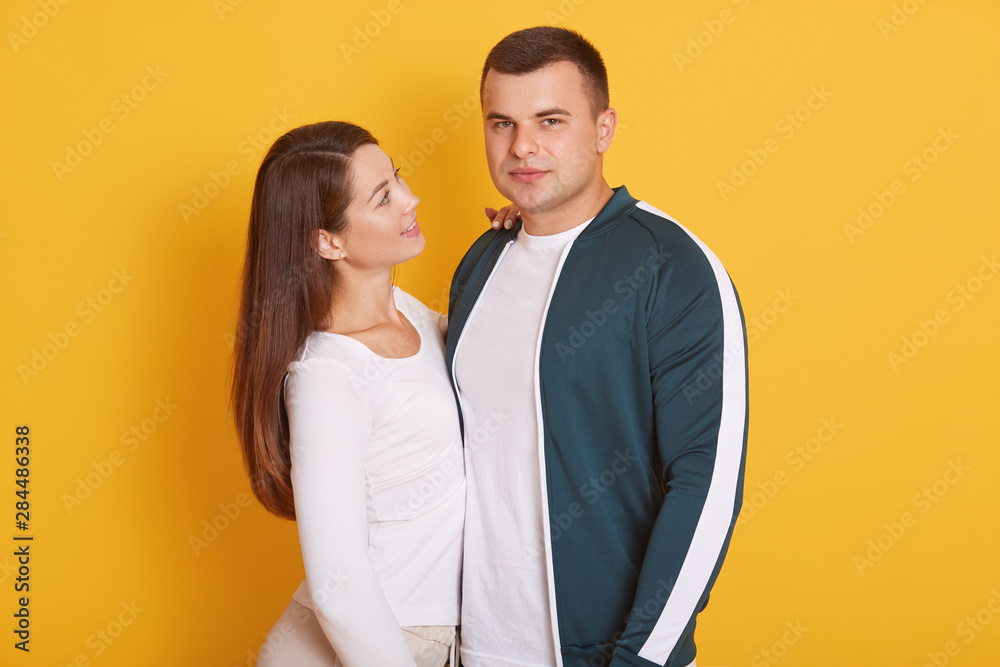 Image of attractive couple, charming woman smiling and looking at her boyfriend, handsome male looking directly at camera, posing isolated over yellow background. Relationship and love concept. Image of attractive couple, charming woman smiling and looking at her boyfriend, handsome male looking directly at camera, posing isolated over yellow background. Relationship and love concept.